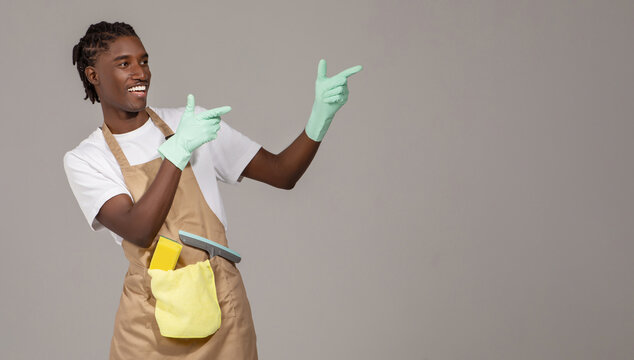 Cheerful young black male cleaner in apron pointing aside at copy space