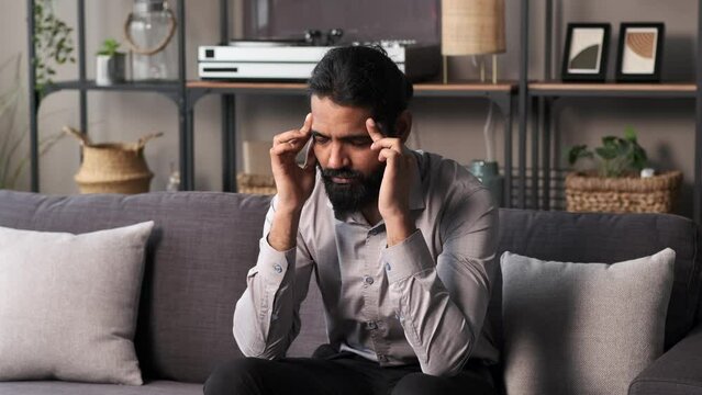 Indian businessman, seated on a sofa in the living room, grapples with a headache, perhaps from the pressures of work. Expressions convey discomfort and introspection.