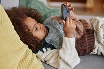 Medium shot of little African American girl in sweater lying on couch at home and playing games...