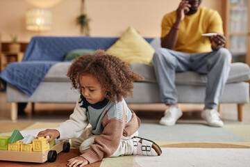 Full length shot of curly-haired little African American girl playing with toy blocks while sitting...