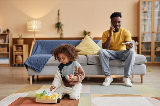 Wide shot of Black father talking on phone holding thermometer while staying at home with sick preschool daughter playing with colorful building blocks on floor
