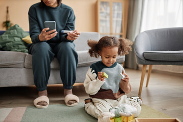 Full shot of African American girl with two ponytails playing with toys on living room floor while unrecognizable mother on sofa holding smartphone and thermometer - obrazy, fototapety, plakaty
