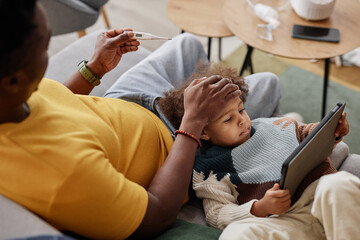High angle view of sick African American girl using tablet with caring father checking her...