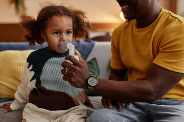 Cropped shot of smiling father putting nebulizer mask on face of unwell African American girl child while sitting on couch at home - Powered by Adobe