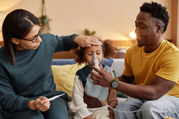Medium long shot of family at home, mom measuring African American daughters temperature while father holding nebulizer mask at childs face - obrazy, fototapety, plakaty