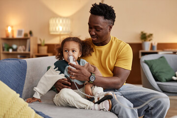 Medium long shot of African American dad embracing little daughter sitting on sofa and holding...