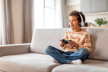 Engrossed black little boy in headphones using game controller at home