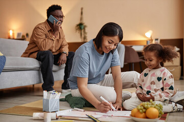 Fototapeta premium Full length shot of cheerful mom mom drawing with daughter sitting on floor together, father using phone in background