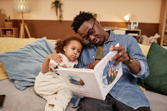 Medium long shot of African American dad wearing glasses reading fairy tales to sleepy little daughter while sitting on couch