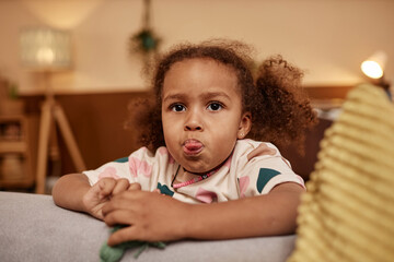 Naklejka premium Medium close up shot of African American preschool girl with curly ponytails sticking out her tongue while leaning on couch