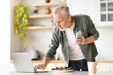 Active senior man browsing his smartphone and using laptop in kitchen