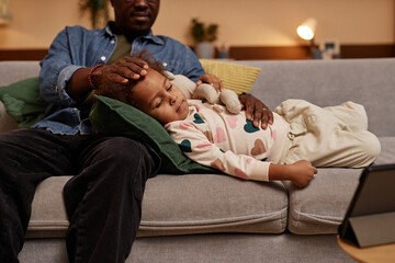 Medium long shot of unwell little African American girl lying on couch watching cartoons on tablet with dad at home
