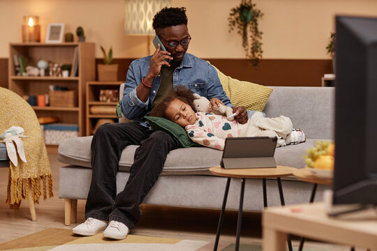Wide shot of sick African American girl lying on couch and hugging toy looking at tablet while her concerned father talking on phone with pediatrician
