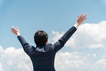 青空に向かって手を広げるビジネスマンの後ろ姿
