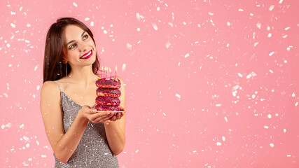 Smiling woman in a sparkling dress admiring a stack of pink doughnuts adorned with birthday