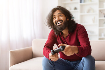 Emotional eastern guy playing video game with joystick at home - obrazy, fototapety, plakaty