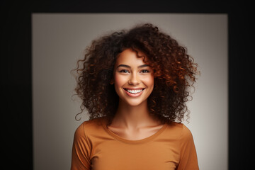 Happy woman with a wide smile and voluminous, curly hair, wearing an ochre-colored top against a neutral grey background.