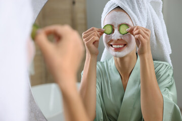 Woman with face mask and cucumber slices near mirror in bathroom. Spa treatments
