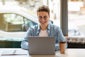 Happy young european guy in glasses using laptop, enjoy remote work or online communication