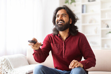 Positive indian guy with remote control in hand watching TV
