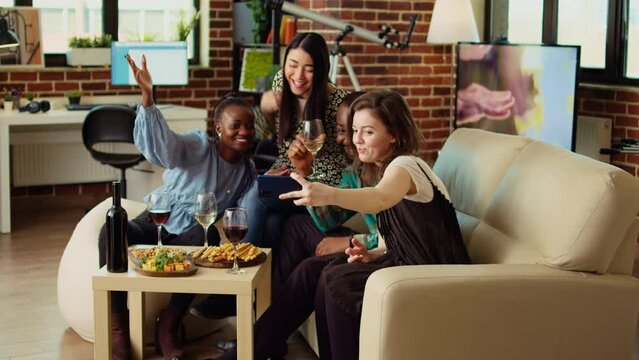 Multiethnic friends taking group pictures with cellphone at apartment gathering. Happy people in living room posing for photo, making funny faces and drinking alcoholic beverages