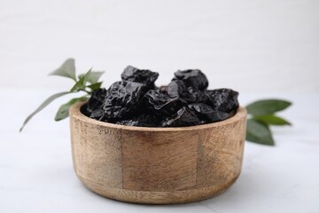 Sweet dried prunes in bowl and green leaves on white table, closeup