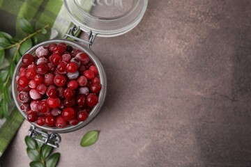 Frozen red cranberries in glass jar and green leaves on brown textured table, top view. Space for text