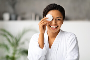 Joyful black woman removing makeup, covering eye with cotton pad