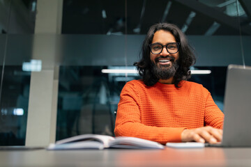 Smiling young indian businessman sitting at worktable at modern office