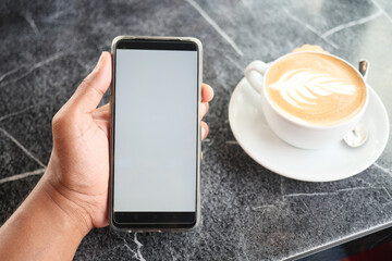  holding a mart phone with white screen and cup of coffee on table 