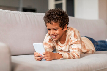 Happy little black boy relaxing with smartphone on couch at home