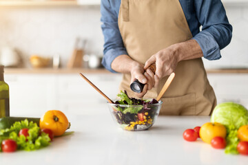 Man in apron seasoning bowl of salad with fresh vegetables in kitchen