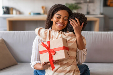 Joyful black mother holding wrapped gift box and affectionately embracing her son