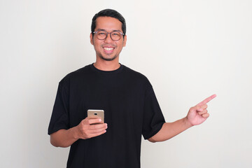 Adult Asian man smiling while holding phone with one hand pointing to the left side