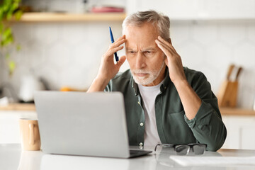 Portrait of stressed senior man using laptop in kitchen interior - Powered by Adobe
