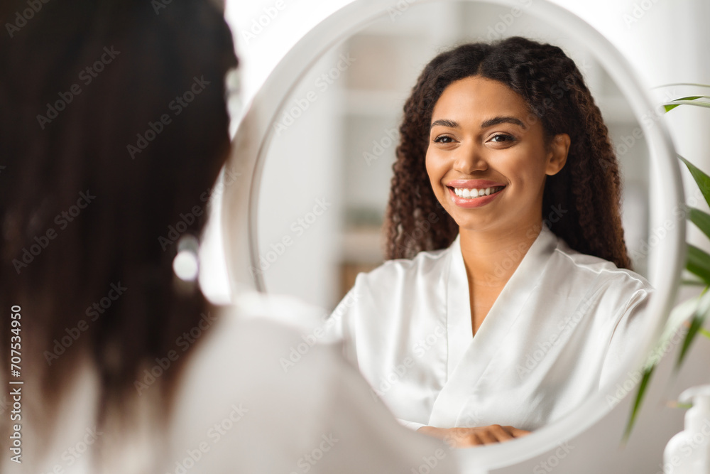 Wall mural Attractive Young Black Woman Wearing Silk Robe Looking In Mirror At Home