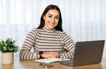 A cheerful young woman with a smartwatch sits at her desk with a laptop