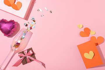 A chocolate bar, paper hearts, blank card, a heart balloon and a clear wine glass on a pink background. Empty space with a flat top-down view.