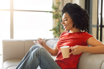 Smiling young black woman writing in notepad and holding mug of coffee