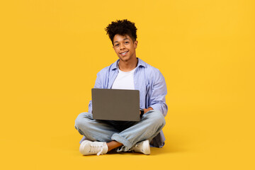 Smiling black male student with laptop sitting cross-legged on yellow background