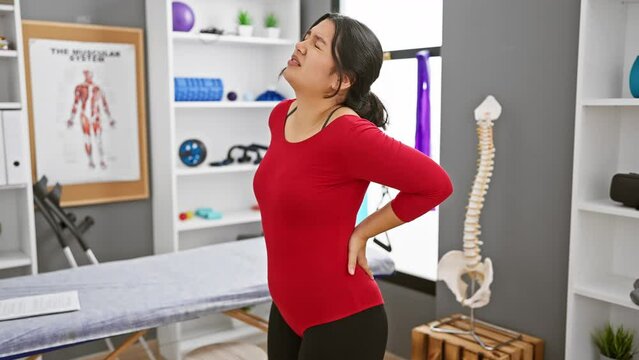 Hispanic woman experiencing back pain in a rehab clinic interior with anatomical charts visible.