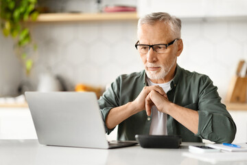 Thoughtful elderly man wearing glasses using laptop and calculator at home