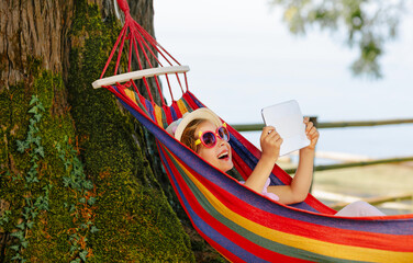 A child swings in a bright hammock outdoors. A child consumes entertainment content from gadgets in...