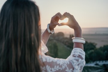 Happy woman in white boho dress making heart sign with hands on sunset in mountains. Romantic woman with long hair standing with her back on the sunset in nature in summer with open hands.
