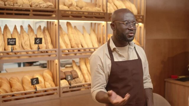 Medium shot of young Black man presenting various types of bread including gluten free baguettes while standing at counter in small bakery and talking with customer