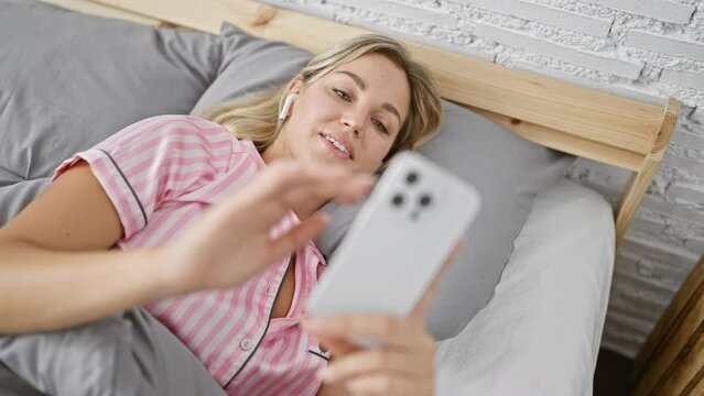 A young blonde woman in pajamas using a smartphone in bed at home, depicting relaxation and modern lifestyle