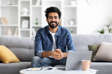 Smiling Indian man wearing glasses sitting at table with laptop at home