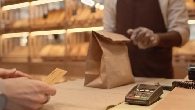 Cropped slowmo of unrecognizable female customer attaching credit card to terminal to make payment at bakery and salesperson in apron giving craft paper bag with her order