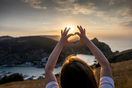 Happy woman on mountain peak, she makes heart shape with hands. Mountain, overlooking sea at sunset. Depicting love shape amidst scenic natural setting.