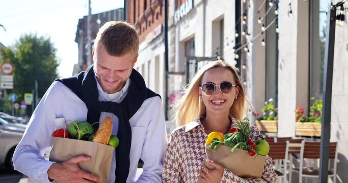 Young family couple walk along city street after grocery shopping carrying paper bags with fresh food. Weekend purchases and happy buyers satisfied with goods